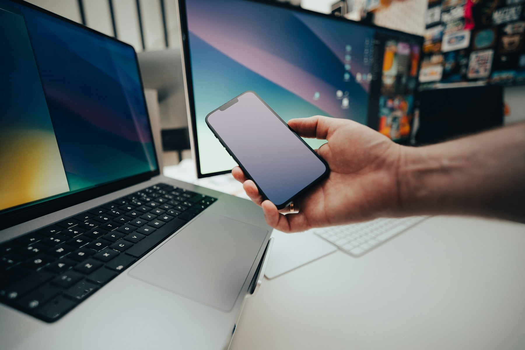 A person checking a smartphone beside a laptop and desktop monitor